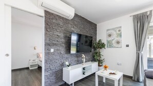 A modern living room with a stone accent wall, a flat-screen TV, a white console table, a small table with cups and a plant, and a grey curtain. - Beach front home in Peroj with house sea view (Peroj)