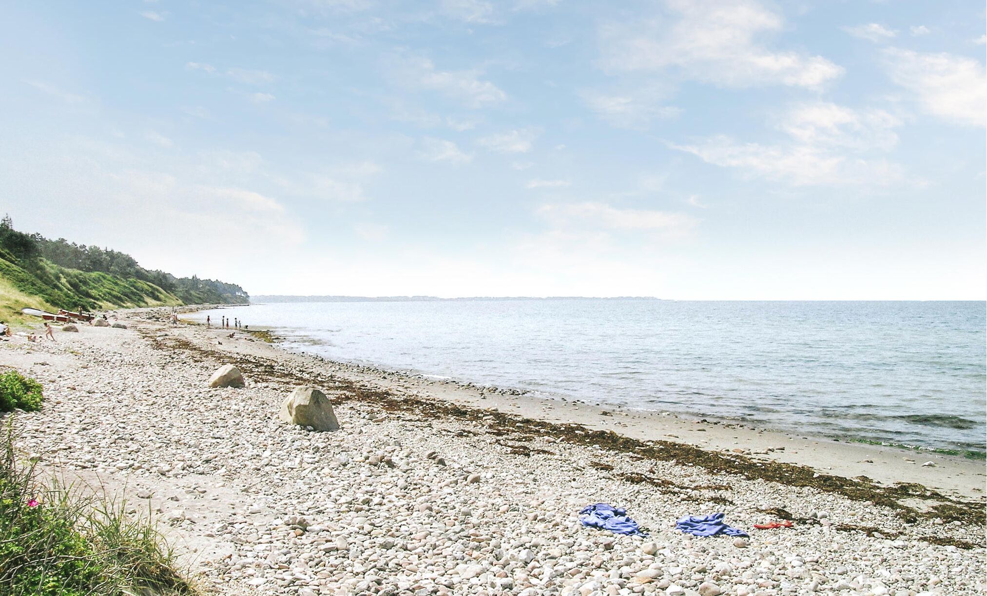 A pebble beach with a calm sea, a distant shoreline, and a grassy area with trees.