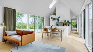 A modern living room with a leather sofa, a dining area with wooden chairs, and a kitchen with white cabinets and a green hanging light. - Awesome home in Fårevejle with kitchen (Fårevejle)