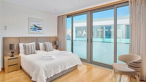 Une chambre moderne avec un grand lit, une table de chevet, une chaise et une vue sur la ville grâce à de grandes fenêtres.