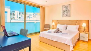 Ein Hotelzimmer mit einem großen Bett, einem Schreibtisch mit einem Laptop, einem Sessel und Blick auf Gebäude durch das Fenster.