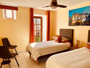 Une chambre d’hôtel avec deux lits, un ventilateur de plafond, une fenêtre avec des rideaux, un tableau représentant un bâtiment et une chaise.