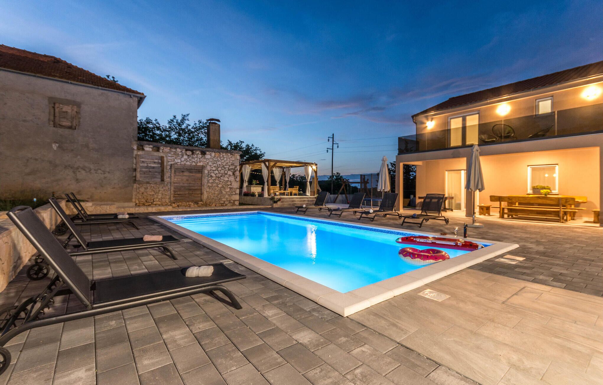 A rectangular swimming pool with lounge chairs and a sunbed, surrounded by stone walls and a modern building with a balcony.