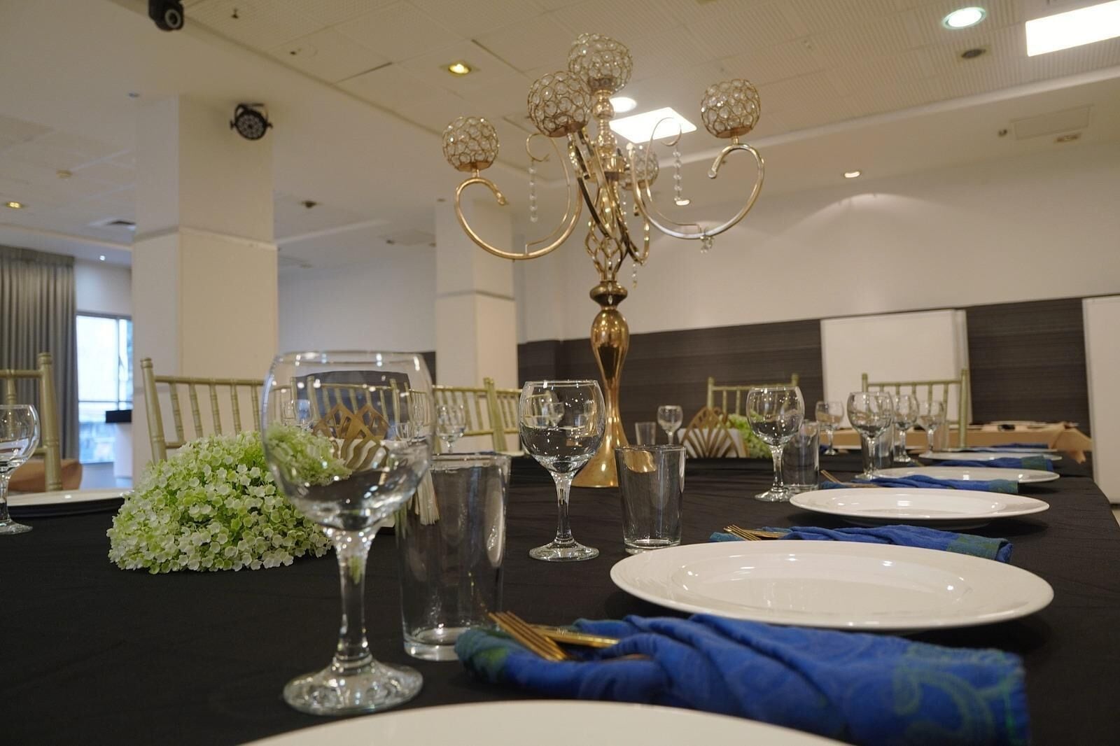 Una mesa de banquete dispuesta para un evento formal, con una lámpara de araña de cristal, platos blancos y copas transparentes.