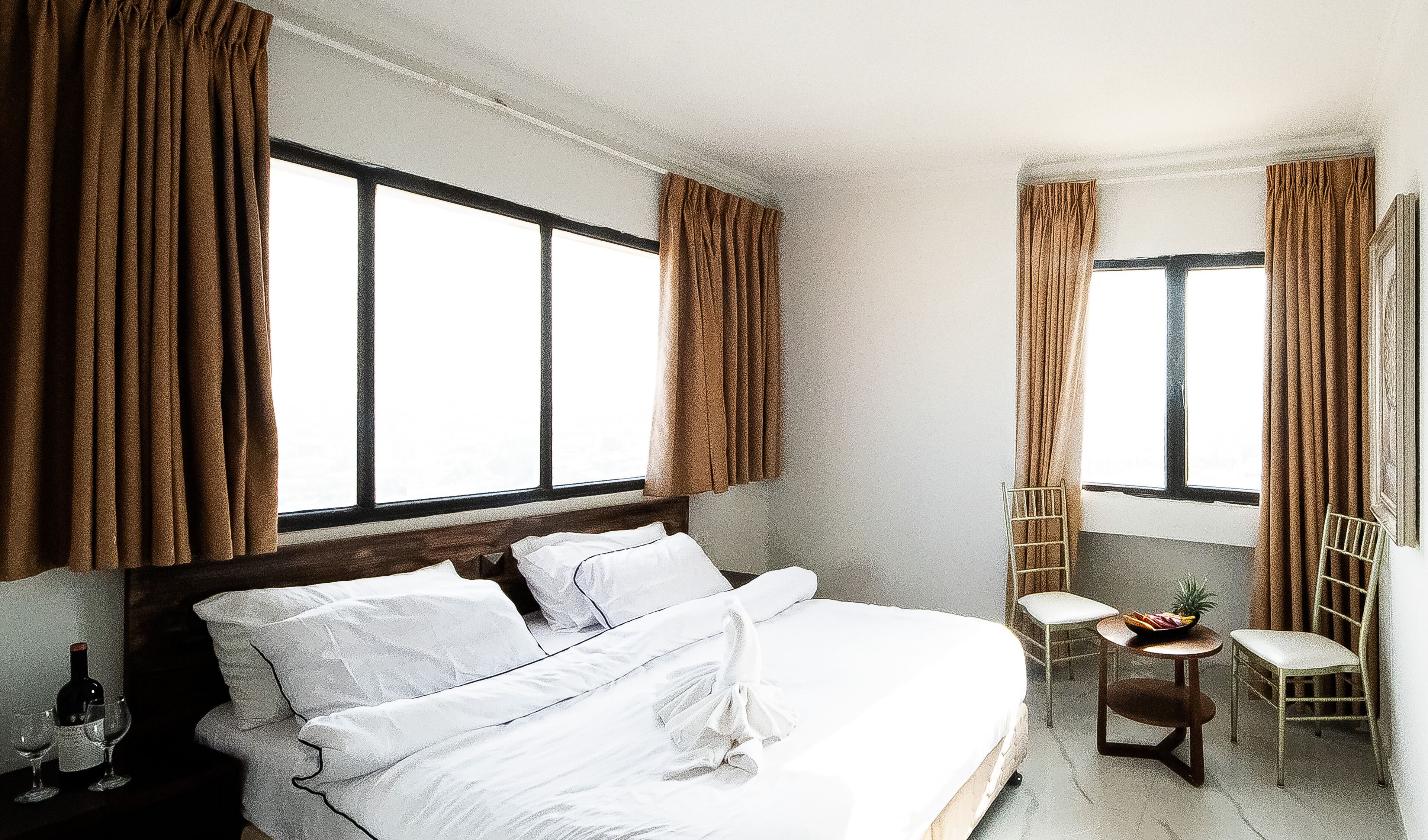 Una habitación de hotel con una cama grande, dos ventanas con cortinas, una mesita con una silla y una botella de vino sobre la mesita de noche.