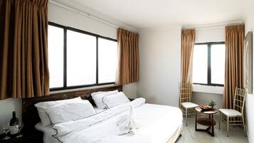 Una habitación de hotel con una cama grande, dos ventanas con cortinas, una mesita con una silla y una botella de vino sobre la mesita de noche.