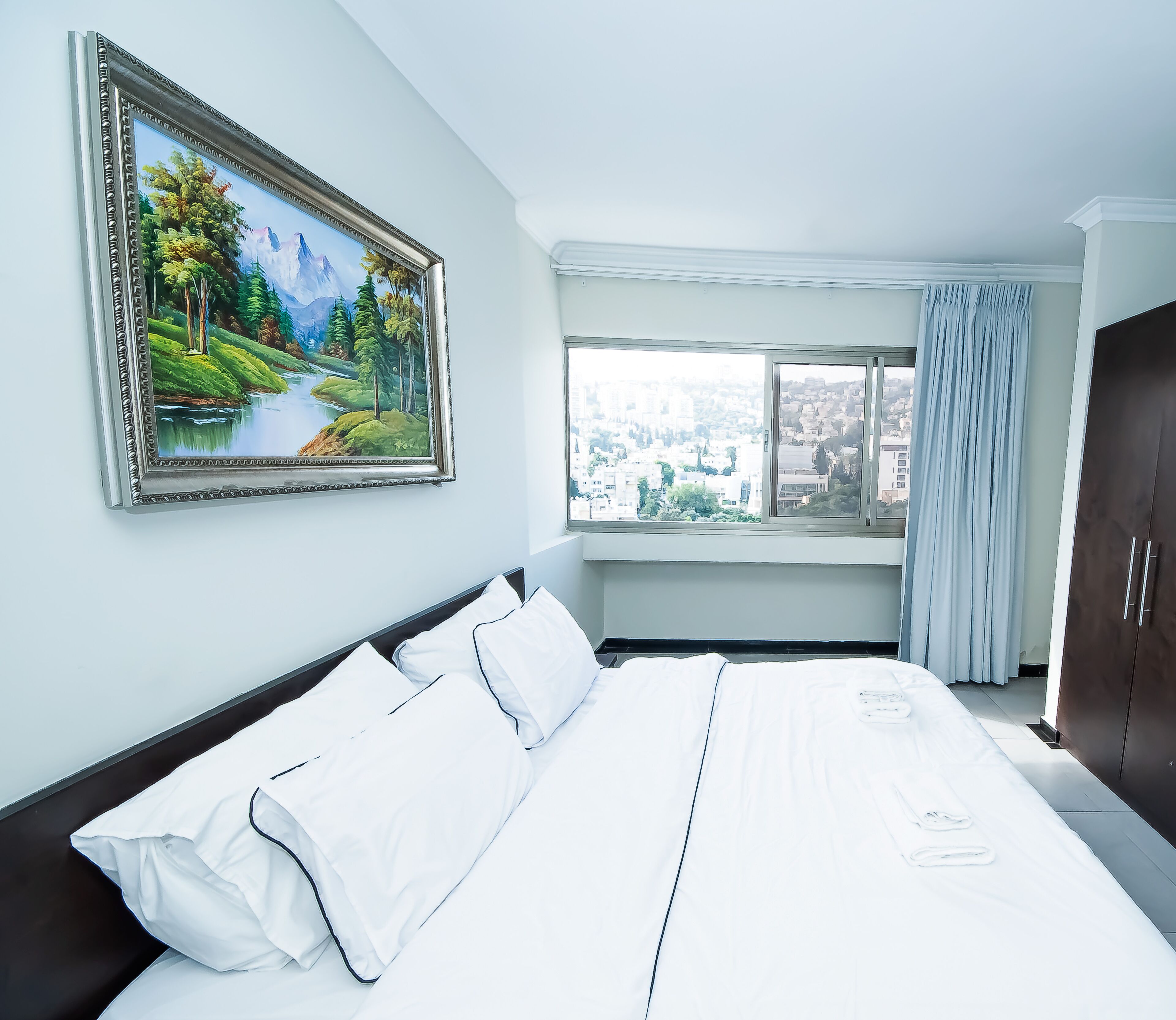 A hotel room with a large bed, a painting of a landscape, a window with a city view, and a wardrobe.