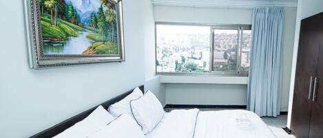 Une chambre d’hôtel avec un grand lit, un tableau représentant un paysage, une fenêtre donnant sur la ville et une armoire.