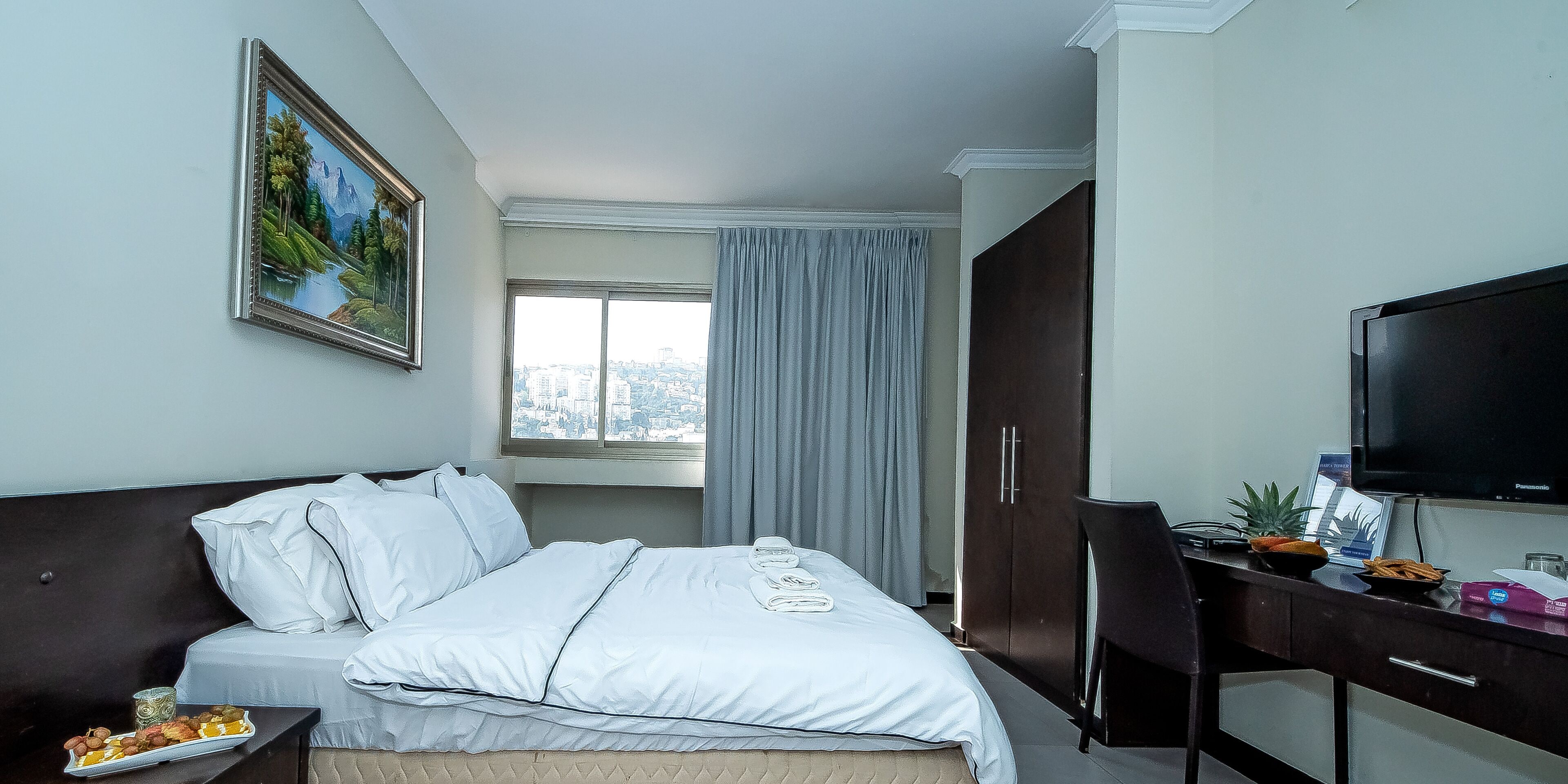 Habitación de hotel con cama, escritorio con computadora y televisión, ventana con vista a la ciudad y un cuadro en la pared.