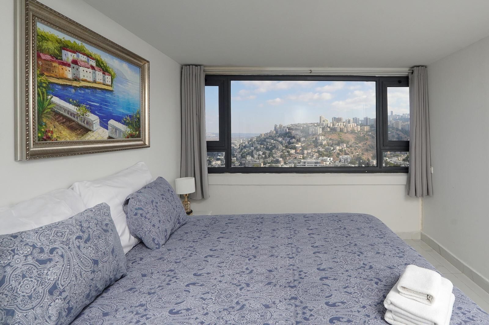 A hotel room with a large bed, a city view, a painting on the wall, and a window with curtains.