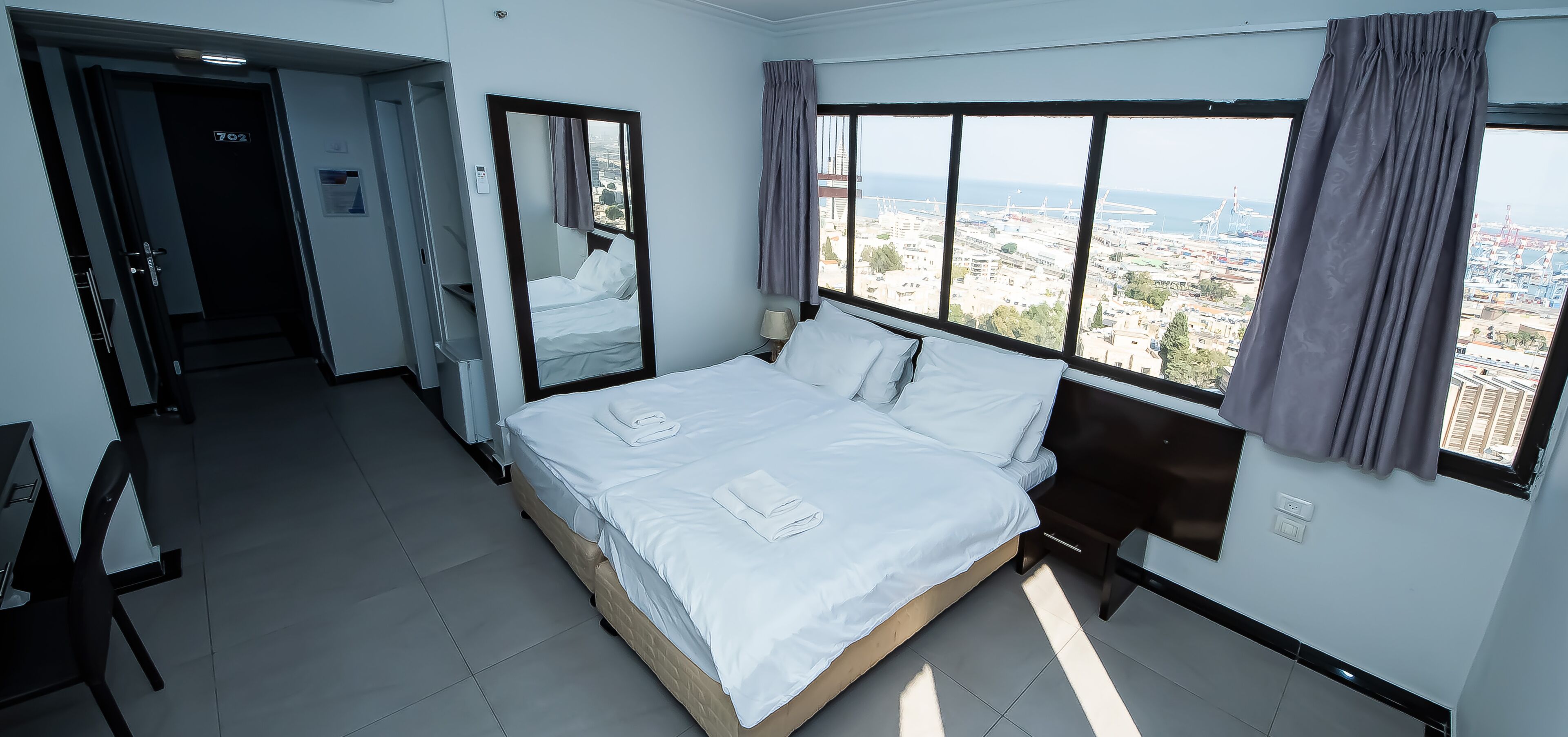 Habitación de hotel con una cama grande, un espejo, una ventana con vistas a la ciudad y una puerta que da a otra habitación.
