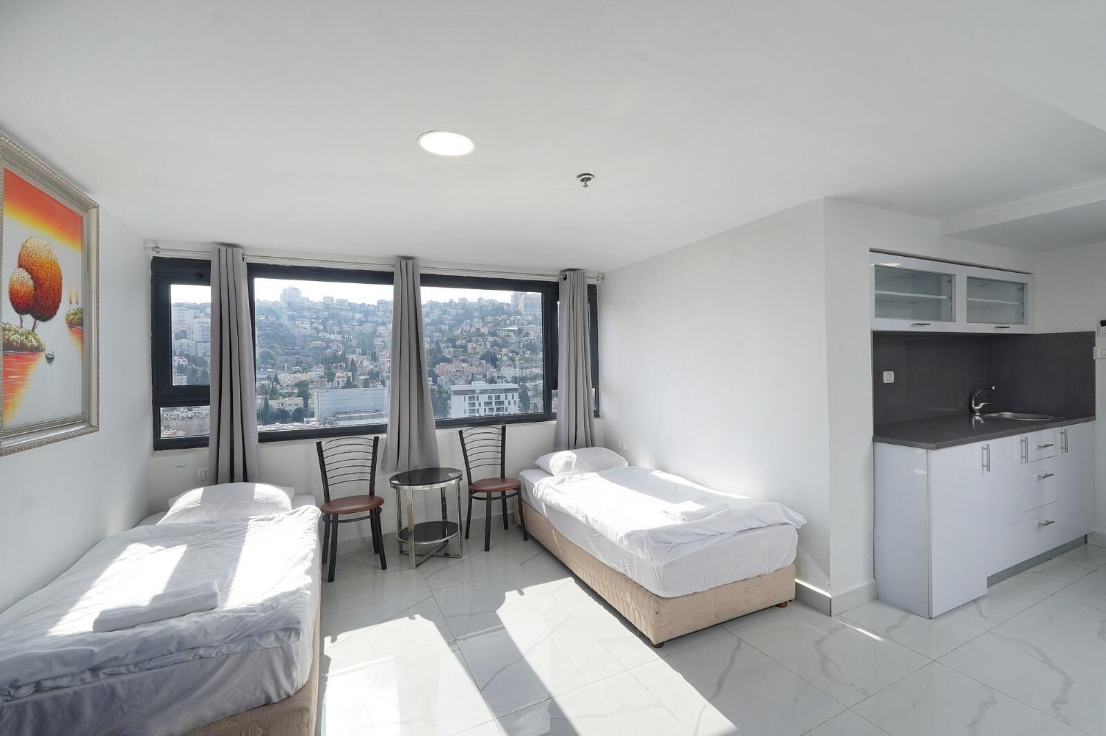 Una habitación de hotel moderna con dos camas individuales, una pequeña mesa de comedor, dos sillas, una zona de cocina y ventanas amplias con vistas a la ciudad.