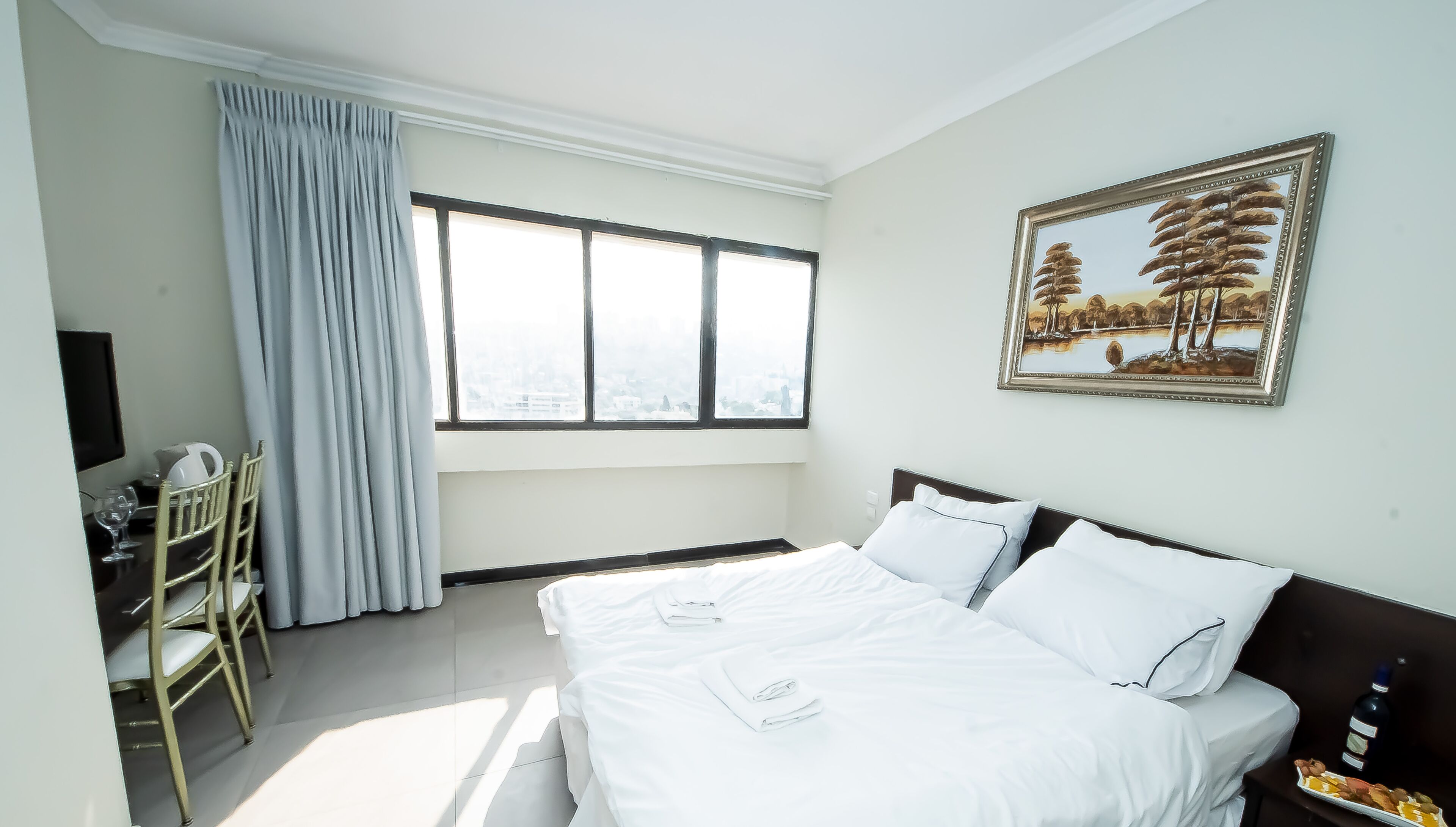 A hotel room with a bed, a desk with a chair, a TV, and a painting on the wall.