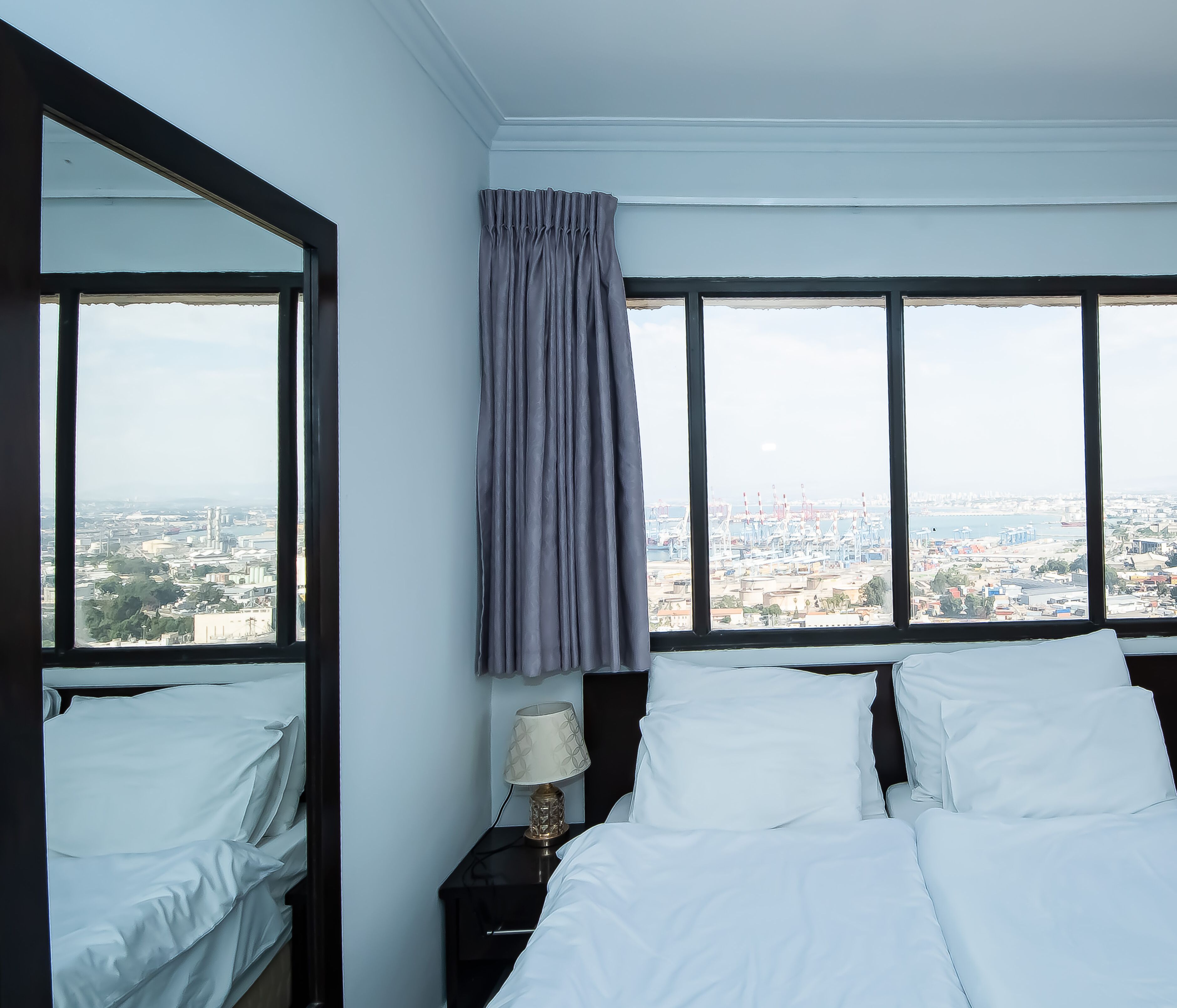 A hotel room with a large bed, a nightstand with a lamp, and a view of a cityscape through large windows.
