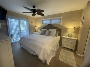 A bedroom with a bed, ceiling fan, bedside lamps, and a view of the outside.