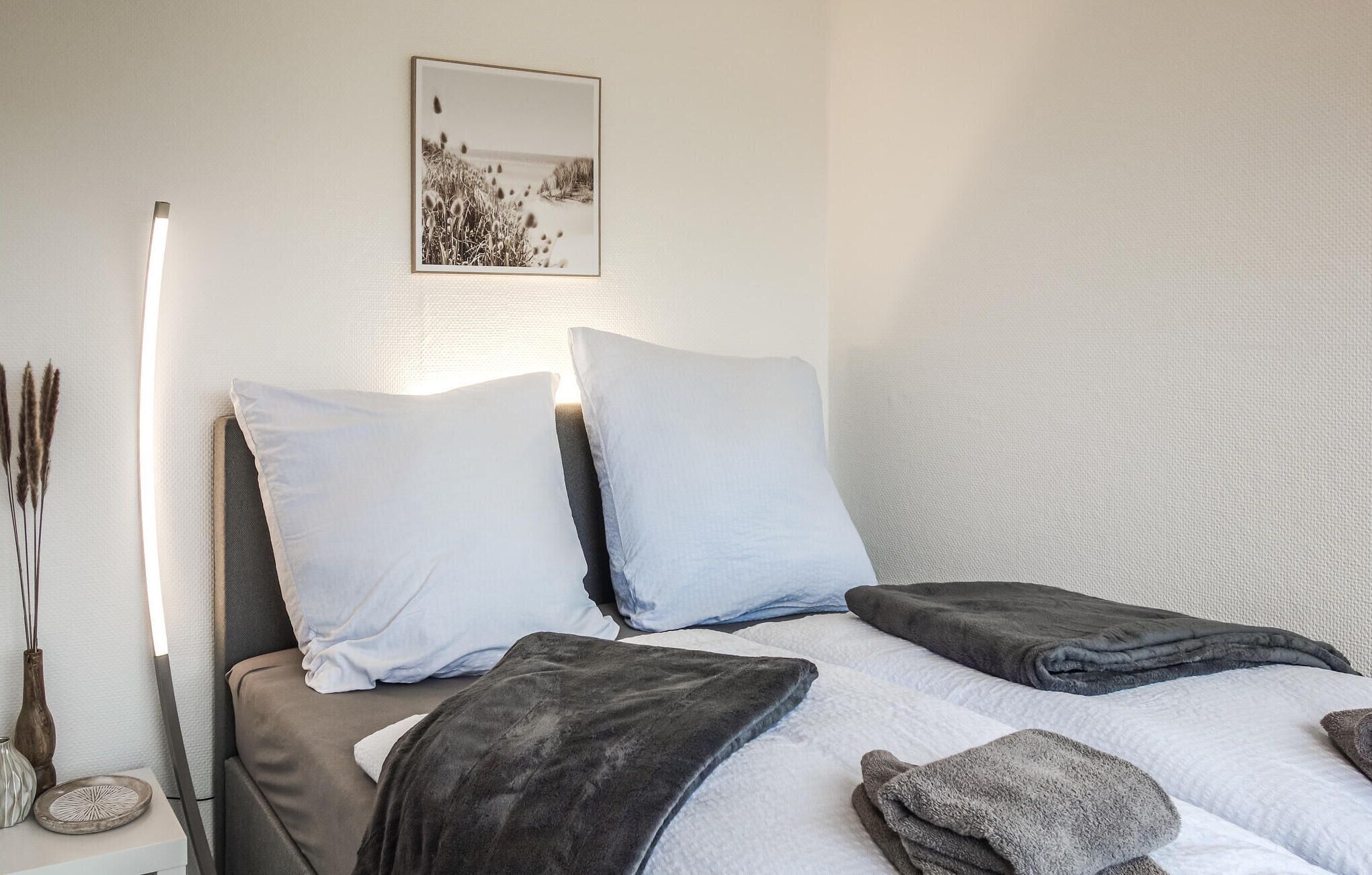 Een net opgemaakt bed met twee kussens en een deken, een nachtkastje met decoratieve objecten, en een lijst met een foto aan de muur.