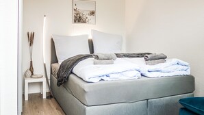 Una cama bien hecha con colchón gris, almohadas blancas y toallas dobladas. Hay una mesita de noche con un florero y una lámpara. Encima de la cama, en la pared, cuelga un cuadro enmarcado.