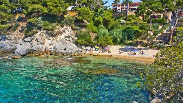A coastal beach with clear water, rocky shore, and buildings in the background. - Beautiful Apartment, Swimming Pool and Beach 2 (Platja d'Aro)