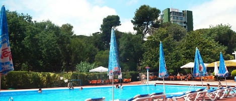 Une piscine entourée de chaises longues et de parasols, le tout bordé de verdure, avec un bâtiment visible au loin.