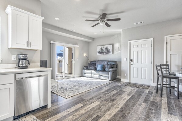 A modern kitchen with white cabinets, a dishwasher, and a ceiling fan. There is a gray rug on the floor and a gray sofa in the living area.