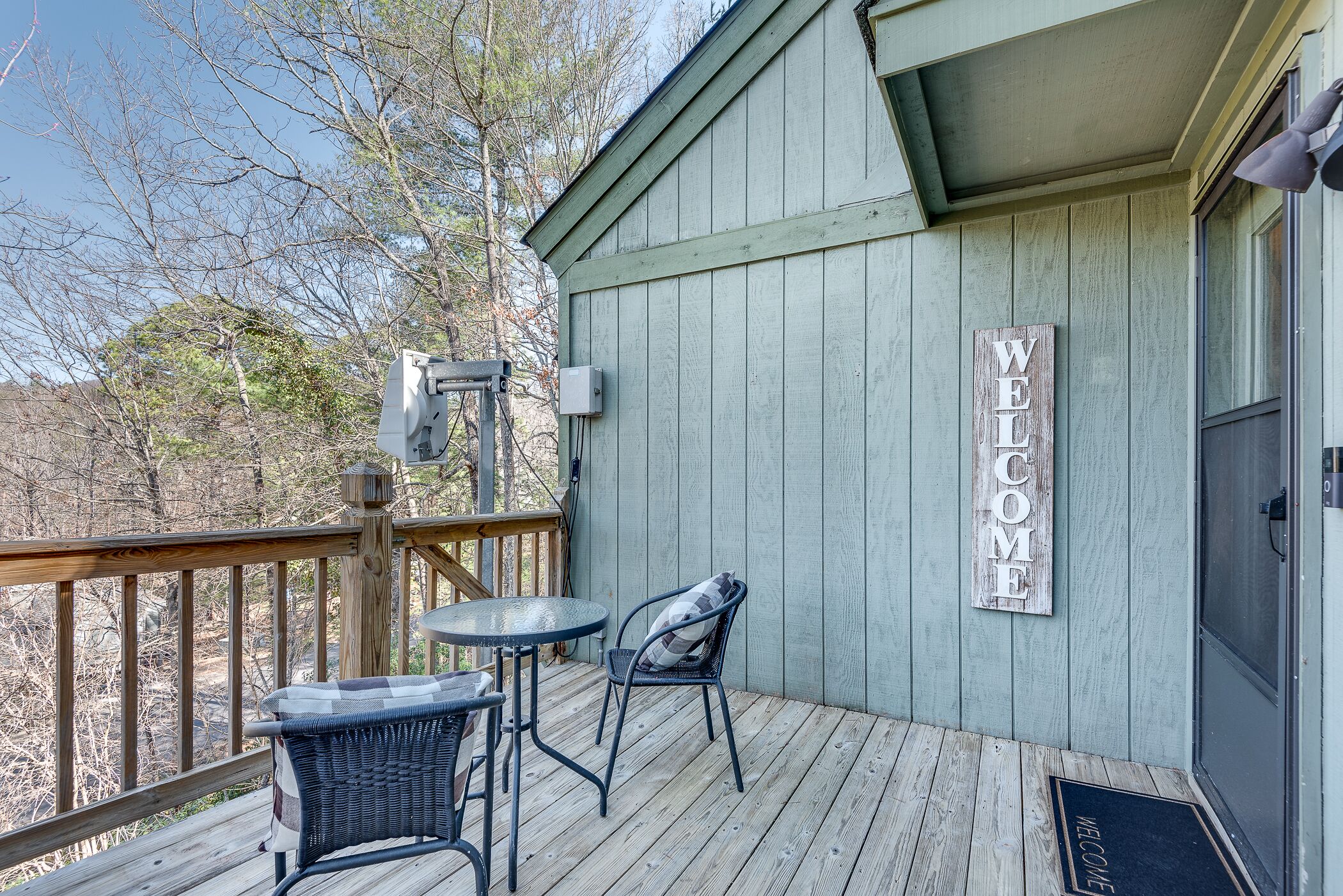 Une terrasse en bois avec deux chaises et une table, un panneau d’accueil et un coffret électrique fixé au mur.