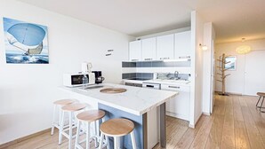 A modern kitchen with white cabinets, a central island with stools, and a painting of a sailboat. - Le Poisson Bleu - by Winkyhouse (Cassis)
