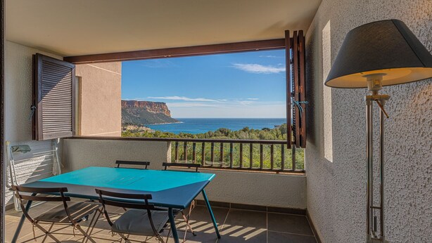 A balcony with a table and chairs, a lamp, and a view of the sea and cliffs. - Le Poisson Bleu - by Winkyhouse (Cassis)