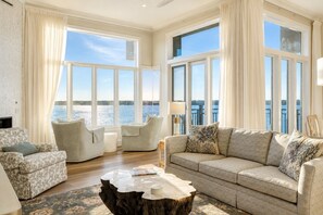 Amplia sala de estar con ventanas grandes, un conjunto de sofás, una mesa de centro y vista al agua.