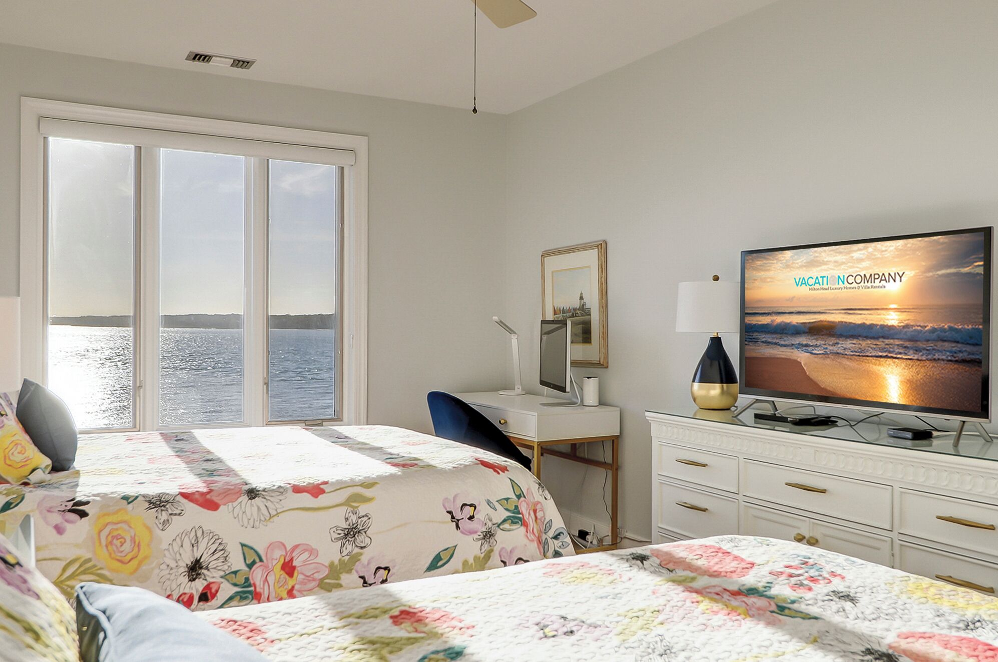 Une chambre d’hôtel avec un lit, un bureau, un téléviseur diffusant une publicité pour une agence de voyages, et une vue sur la mer par la fenêtre.