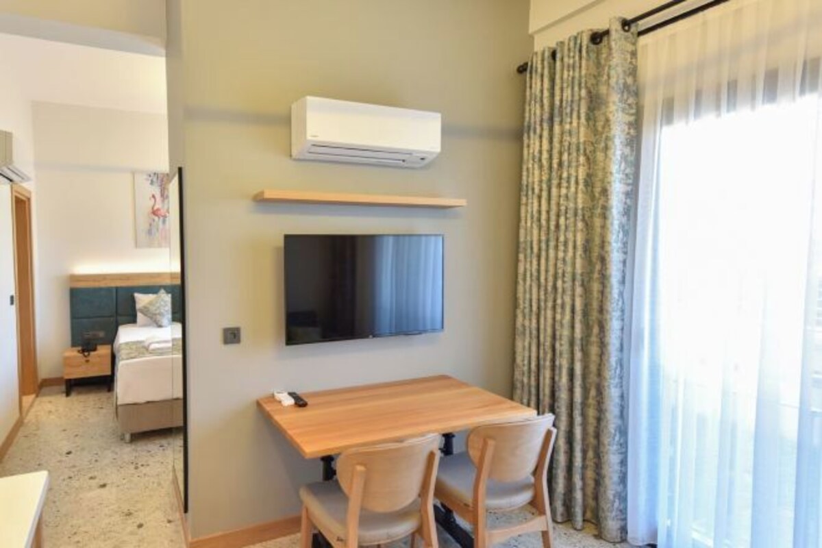 A hotel room with a bed, a TV mounted on the wall, a wooden table with two chairs, and an air conditioning unit.