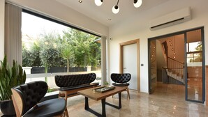 A modern living room with a sofa, chairs, a coffee table, and a large window offering a view of trees and a building. - Avilia Suites (Fethiye)