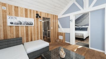 A room with a bed, a sofa, a table with a decorative item, wooden paneling, a door leading to a balcony, and a wall-mounted light fixture.