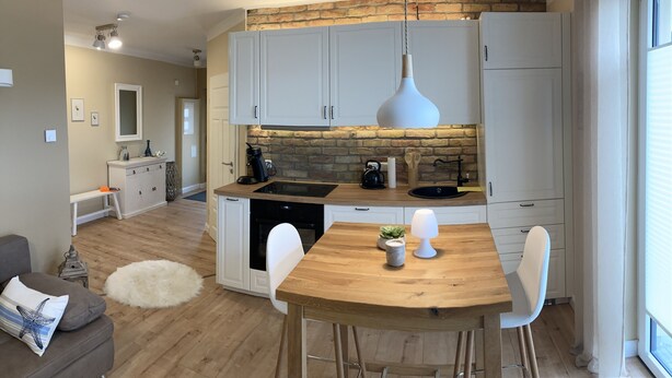 Een moderne keuken met witte kasten, een houten aanrecht en een betegelde spatwand. Er is een eettafel met stoelen en een hangende lamp.