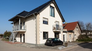 Een twee verdiepingen tellend huis met een auto ervoor, een balkon op de eerste verdieping en een rode dakbedekking.