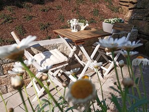 Una mesa de madera y sillas blancas dispuestas en un patio de piedra, con una maceta y un farol.