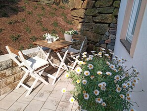 Un patio al aire libre con una mesa de madera y sillas blancas, un muro de piedra y un cantero con margaritas blancas.
