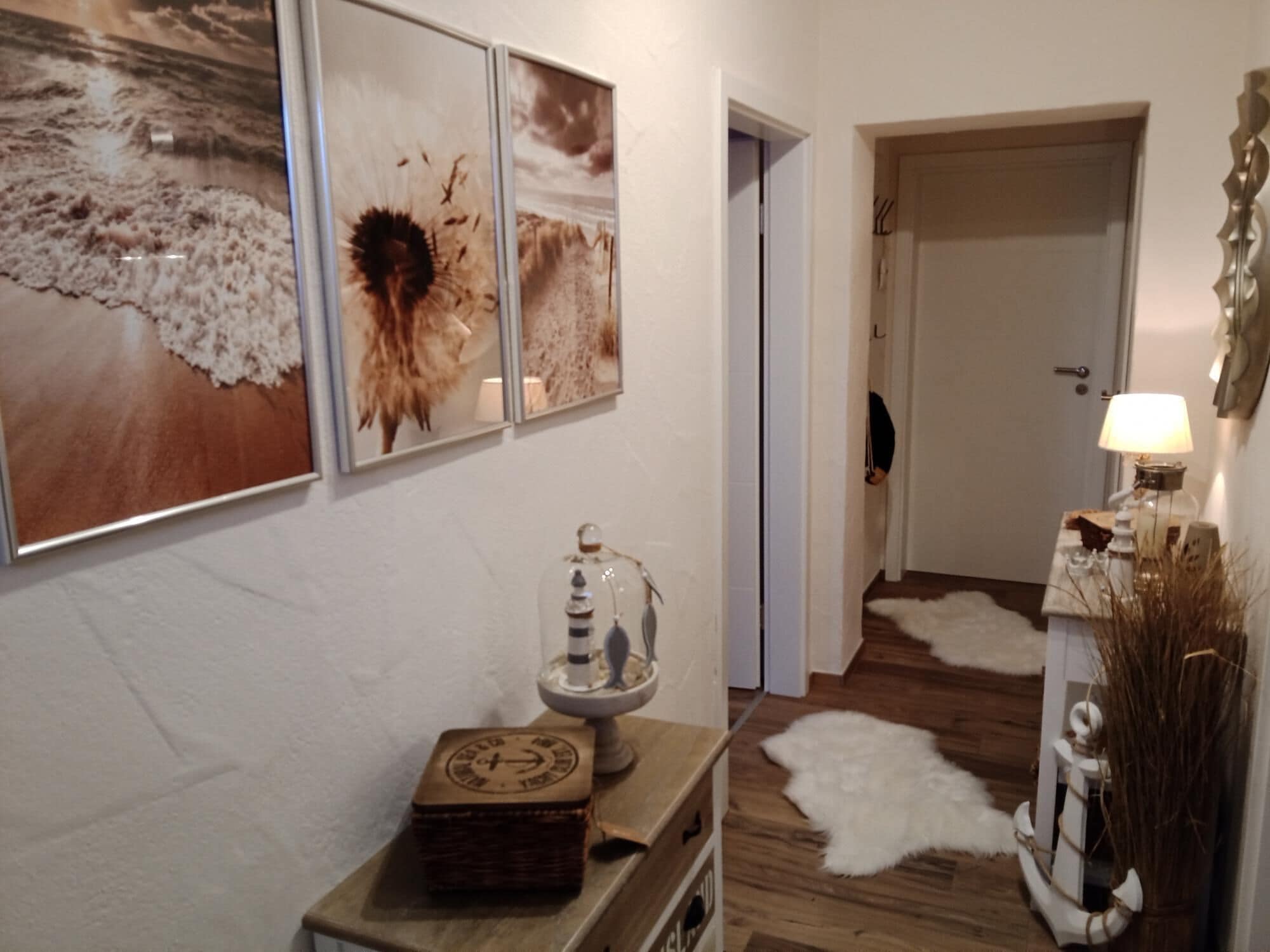 Un couloir doté d’un plancher en bois, d’une porte blanche et d’un mur orné de trois tableaux encadrés. On y trouve une petite table avec une lampe et des objets décoratifs.