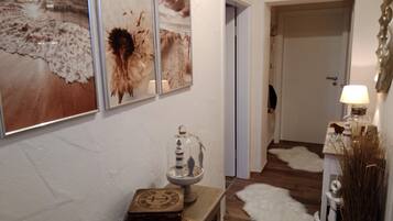 Un couloir doté d’un plancher en bois, d’une porte blanche et d’un mur orné de trois tableaux encadrés. On y trouve une petite table avec une lampe et des objets décoratifs.
