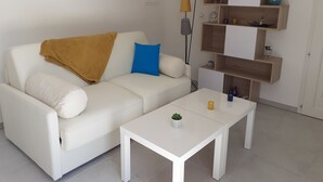 Een witte bank met een bruine plaid en een blauw kussen, een witte salontafel met een kleine plant, en een houten dressoir met diverse objecten.