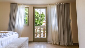 Una habitación con cama, un banco, una ventana con cortinas y un balcón con barandilla.