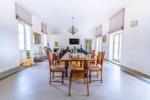 Un comedor espacioso con una gran mesa de madera rodeada por seis sillas, una araña y ventanas amplias con cortinas.