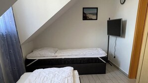 Une petite chambre mansardée de forme triangulaire, avec deux lits, un téléviseur à écran plat et une horloge au mur.