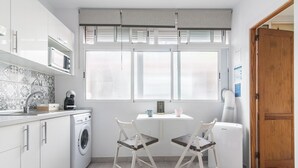 A compact kitchen with a washing machine, two chairs, a small table, and a window with blinds. - Minimalist Studio 3 Minutes Walk to Las Canteras Beach (Las Palmas de Gran Canaria)