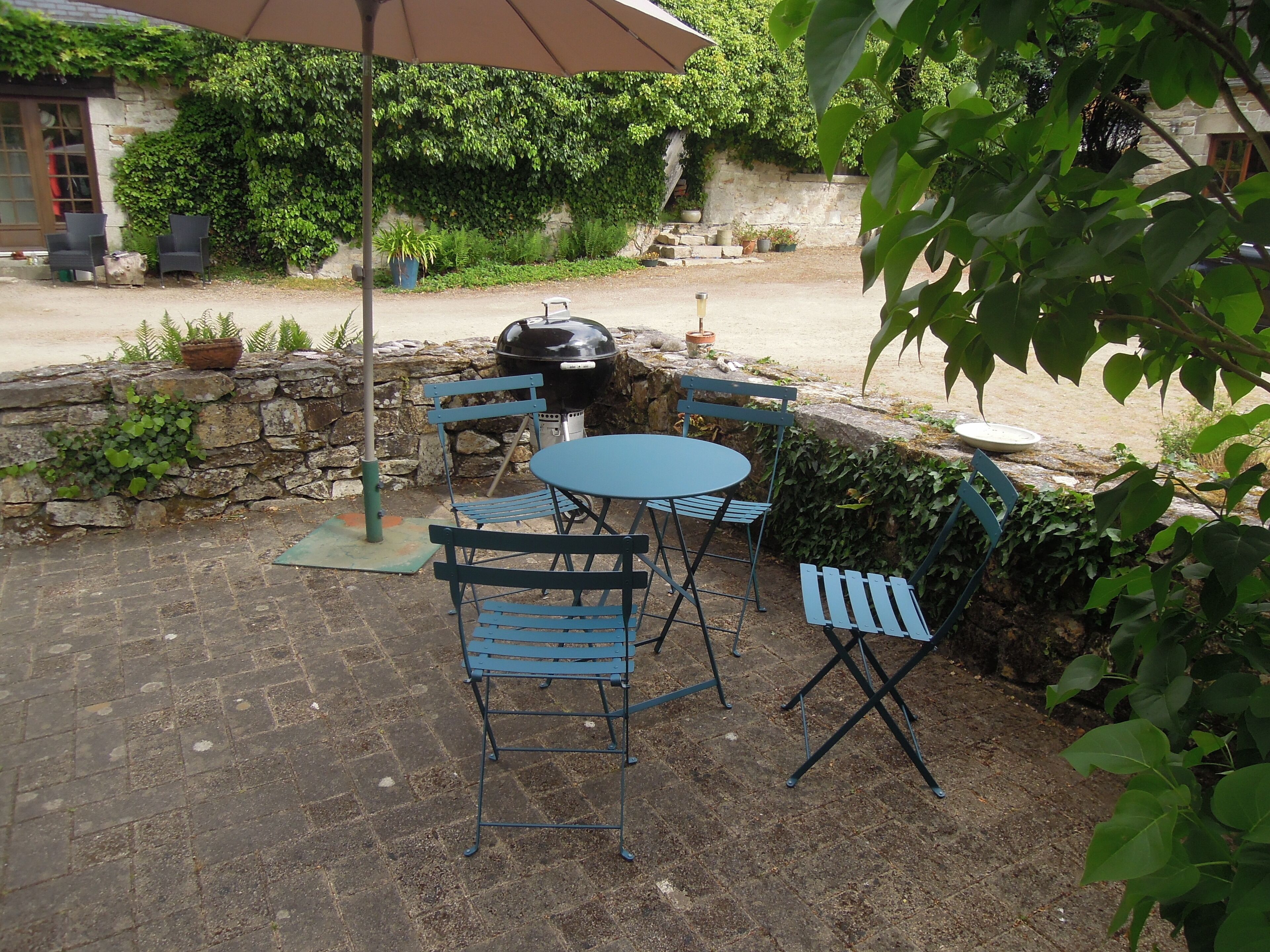 Een terras buiten met een ronde tafel en blauwe stoelen, een barbecue en potplanten.