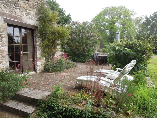 Un bâtiment en pierre doté d’une grande fenêtre et d’une porte, entouré d’une végétation luxuriante et d’un patio dallé aménagé avec du mobilier d’extérieur.