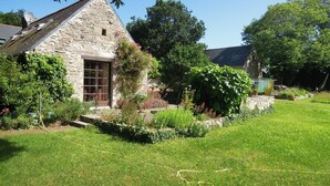 A stone house with a sloped roof, a large window, and a garden with various plants and flowers.