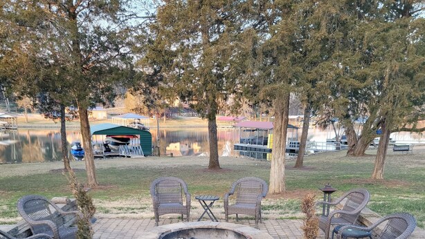 A fire pit surrounded by wicker chairs and a small table, with a paved walkway leading to a lake.