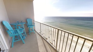 A balcony with two turquoise Adirondack chairs and a small table, overlooking a sandy beach and calm sea.