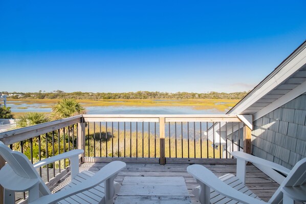 A wooden deck with white chairs and a view of a water body and distant landscape. - Ocean & Wetland views, 3 decks, steps from the beach, 4 BRs, sleeps 10. (Oak Island)
