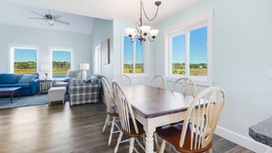 A dining area with a wooden table and chairs, a ceiling fan, and large windows with a view of greenery. - Ocean & Wetland views, 3 decks, steps from the beach, 4 BRs, sleeps 10. (Oak Island)
